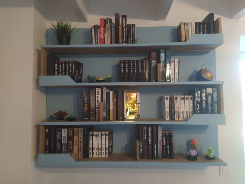 Étagères bleu-bois avec des romans policiers/thrillers (Coben, Thilliez, Chattam, Carrisi, Gardner). Inclut un diorama lumineux et plantes déco.