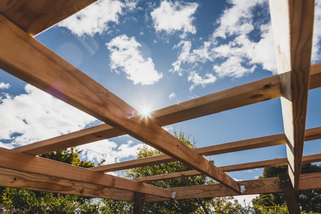 Structure de pergola en bois vue d'en bas, ciel bleu clair avec nuages blancs. Le soleil brille à travers les poutres, créant un éclat lumineux.