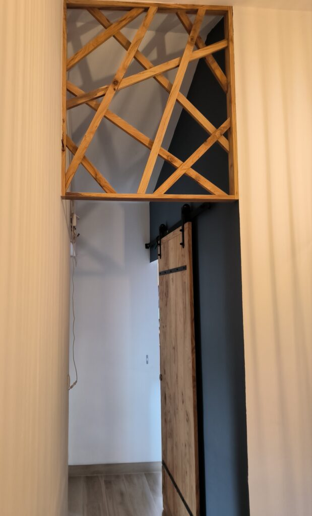 Cloison Déco Bois Moderne et Porte Grange Coulissante Passage entre deux murs blancs, surmonté d'une cloison décorative en bois clair à croisillons, au-dessus d'une porte de grange coulissante rustique.