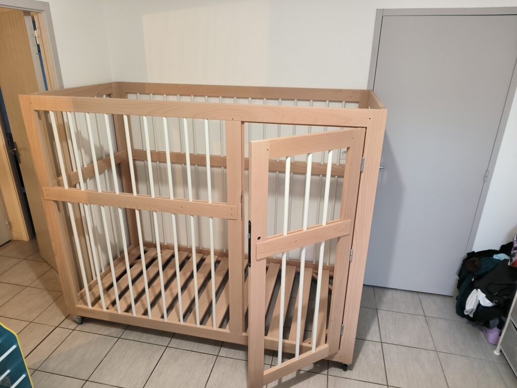 Grand parc carré pour bébé en bois clair et barreaux blancs, monté sur roulettes. La porte d'accès est ouverte, montrant le sommier à lattes.