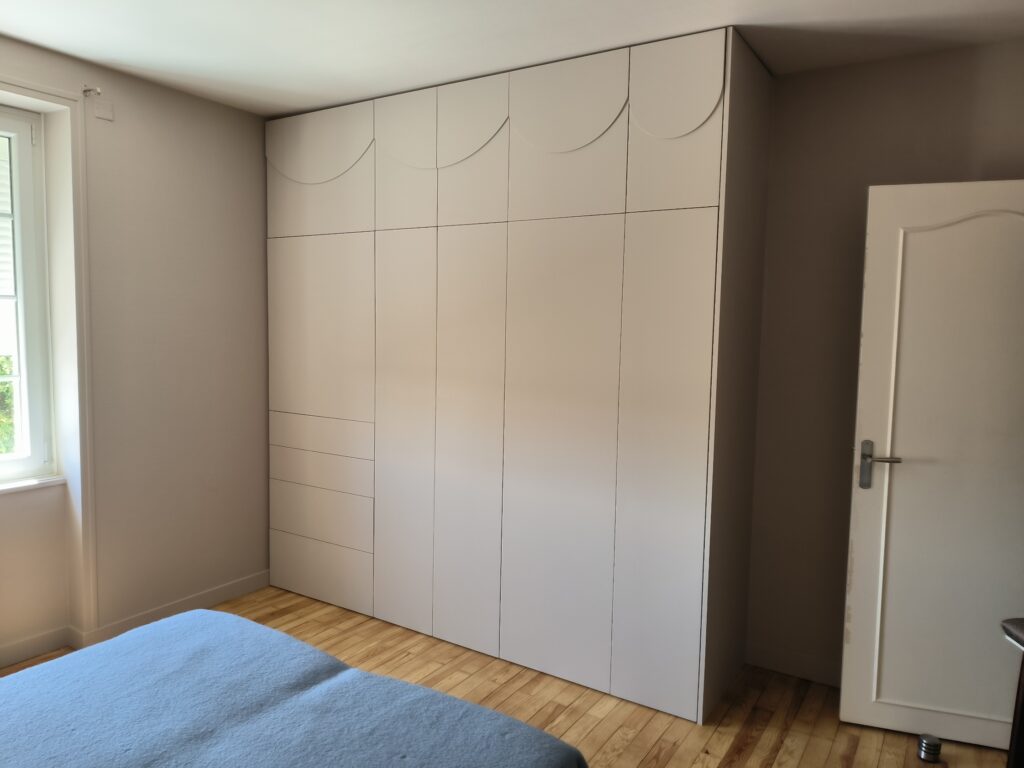 Armoire encastrée sur mesure beige avec motif festonné dans une chambre lumineuse avec parquet et lit bleu.